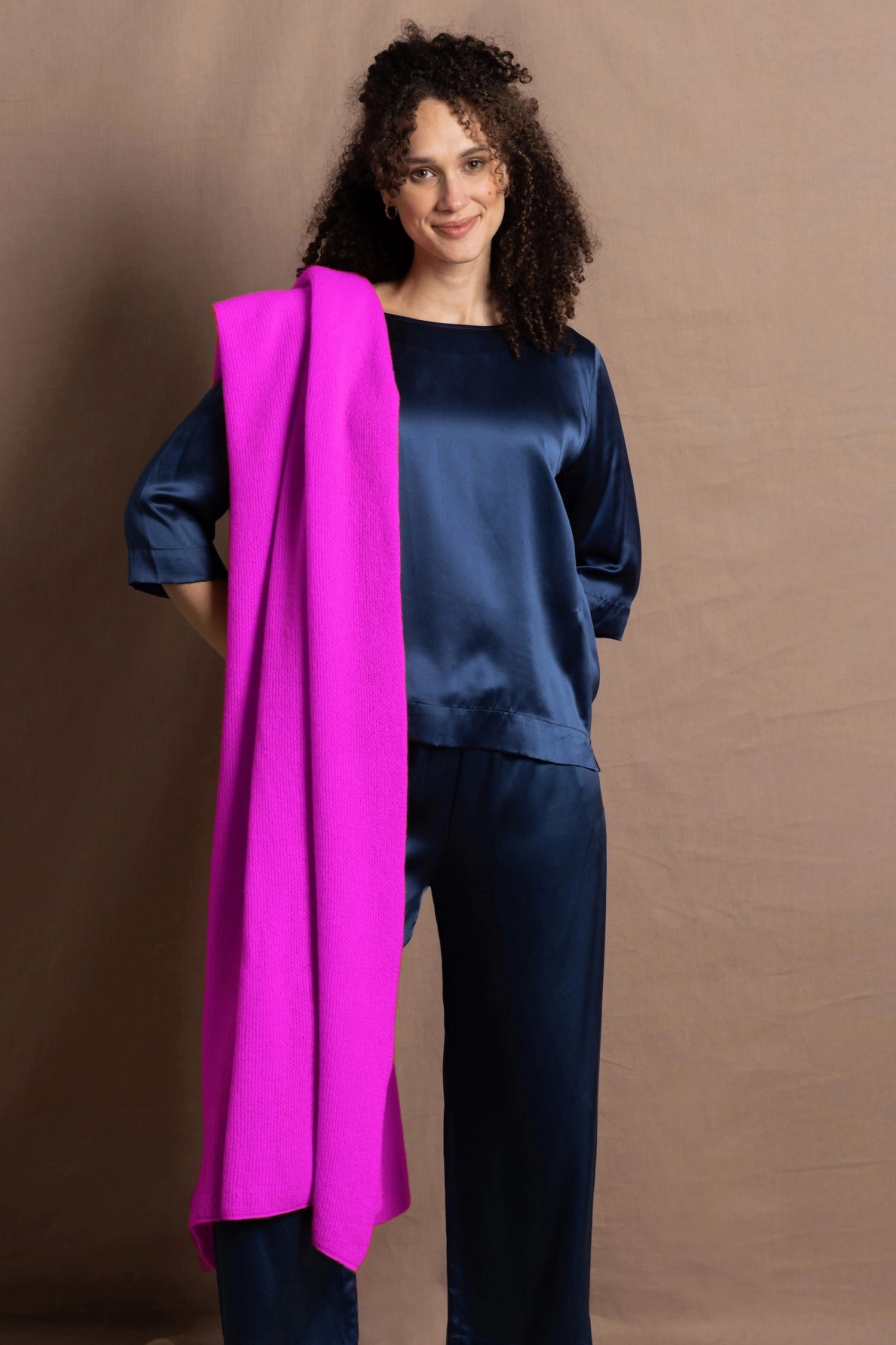 A woman wears a large neon pink cashmere wrap over her shoulder over a full teal silk to and trouser outfit