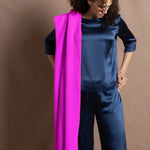 A woman wears a large neon pink cashmere wrap over her shoulder over a full teal silk to and trouser outfit