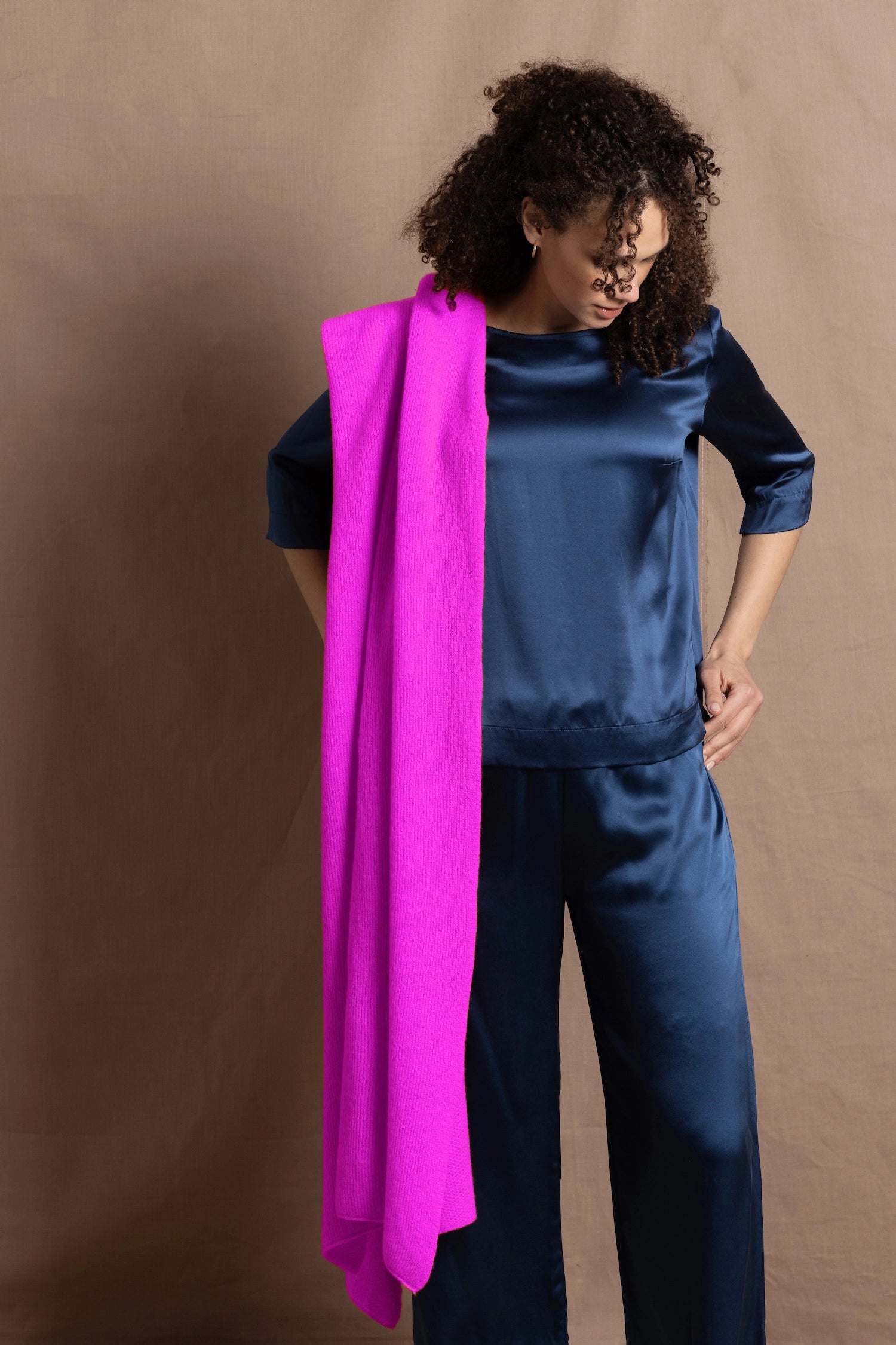 A woman wears a large neon pink cashmere wrap over her shoulder over a full teal silk to and trouser outfit