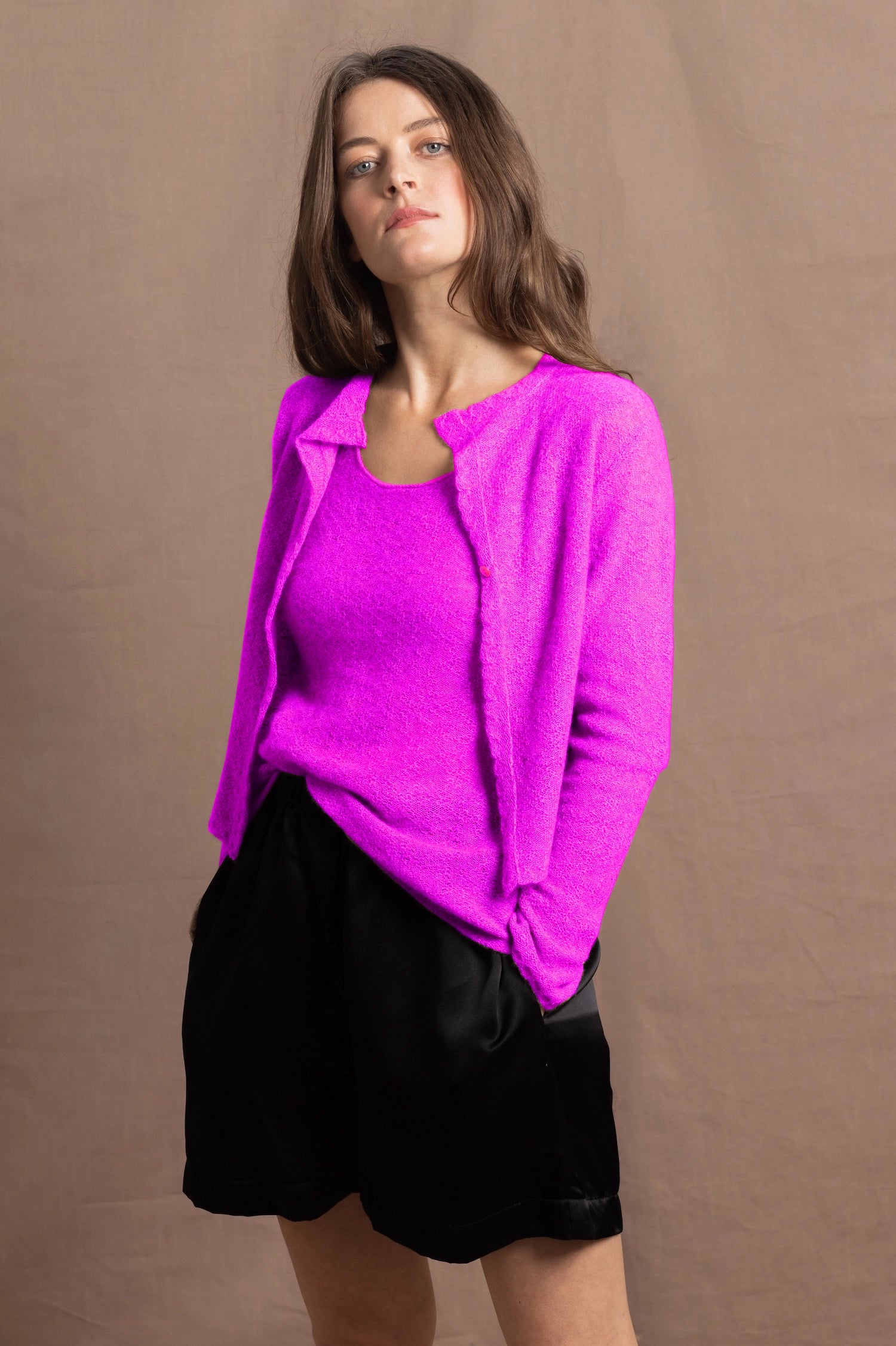 A woman wears a cashmere vest top and matching cardigan in neon pink with black silk shorts