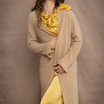 a woman wears a long cashmere cardigan coat in sand beige colour over silk top and trousers in butter yellow by Irish brand Madigan