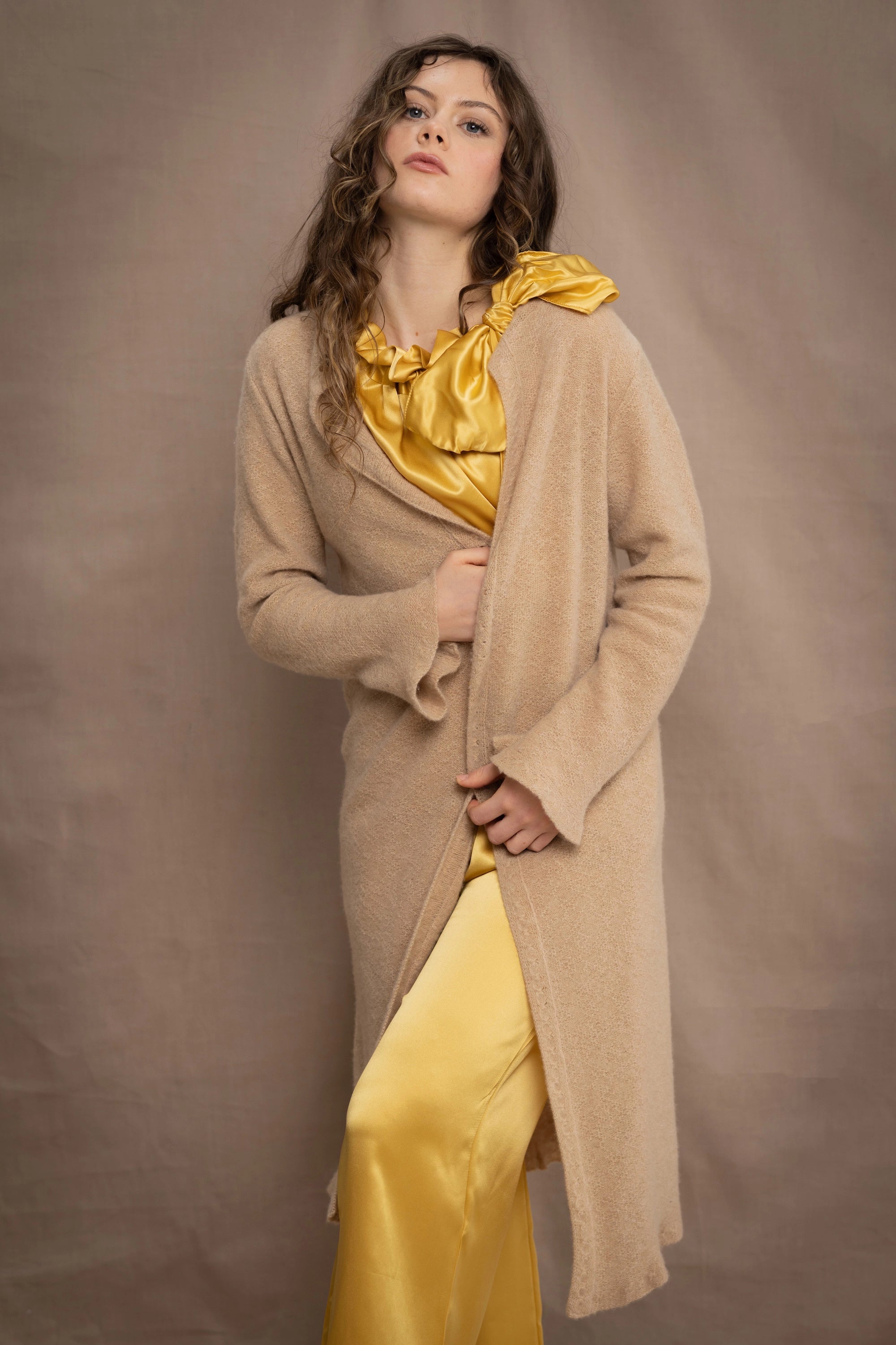 a woman wears a long cashmere cardigan coat in sand beige colour over silk top and trousers in butter yellow by Irish brand Madigan