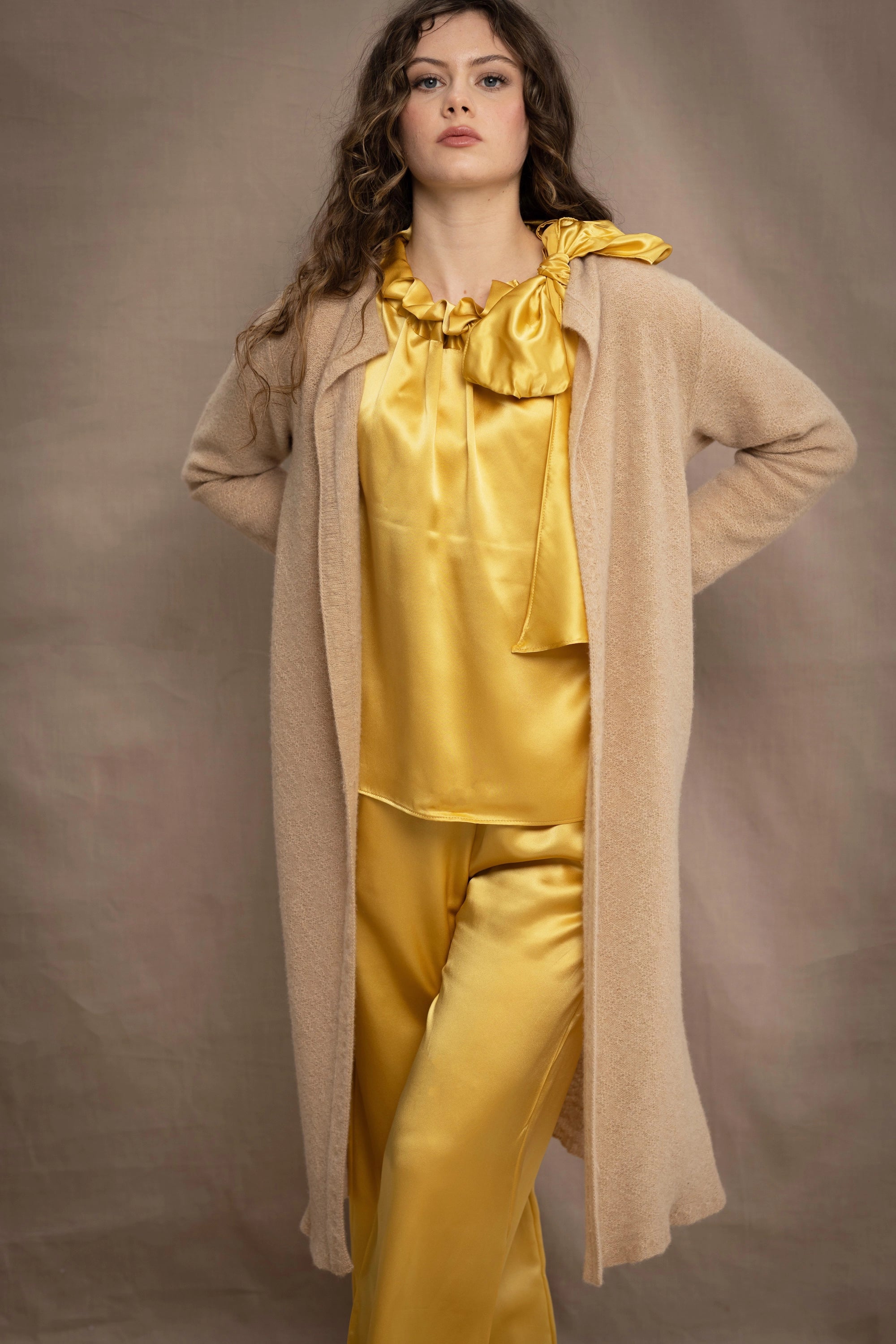 a woman wears a long cashmere cardigan coat in sand beige colour over silk top and trousers in butter yellow by Irish brand Madigan