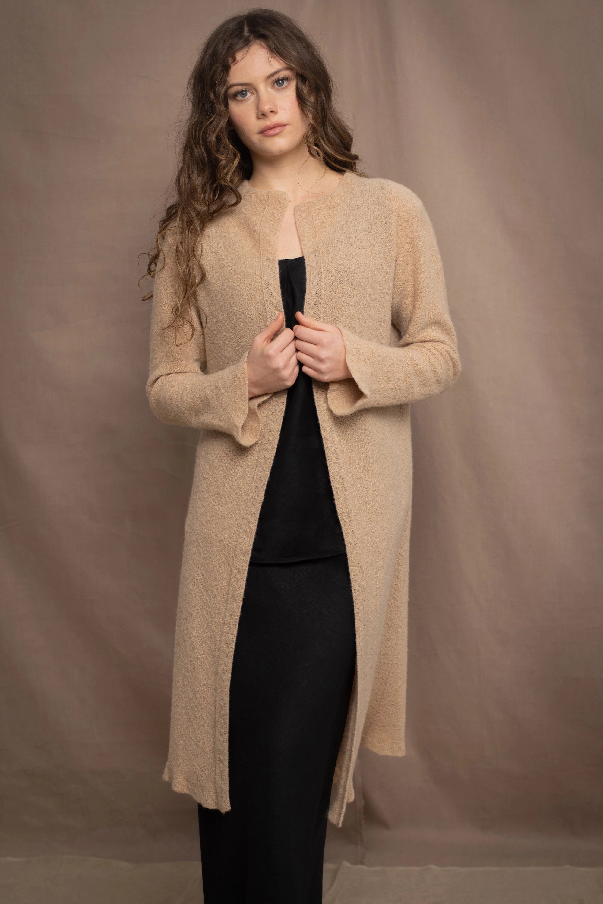 a woman wears a long cashmere cardigan coat in sand beige colour over black linen clothing by Irish brand Madigan