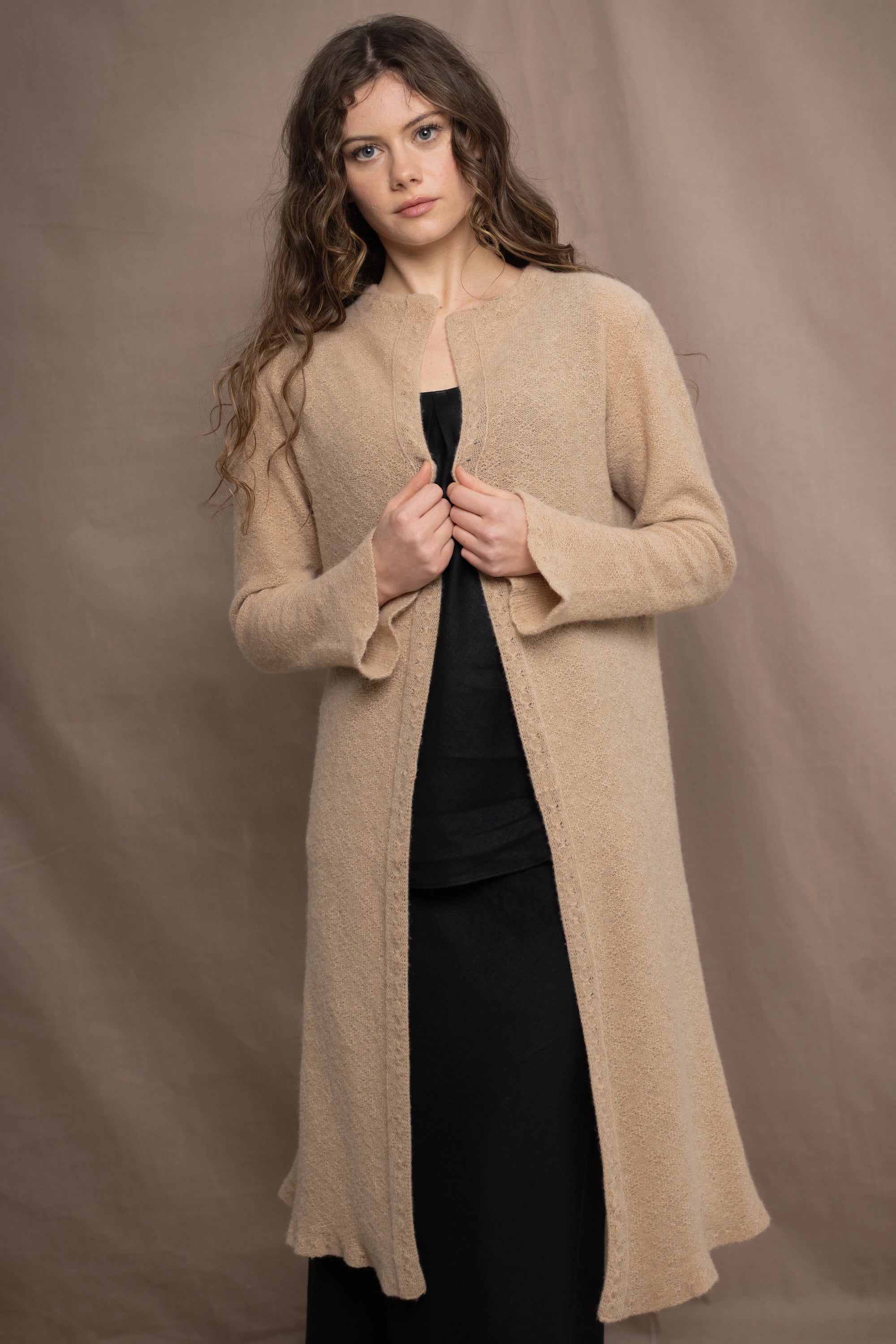 a woman wears a long cashmere cardigan coat in sand beige colour over black linen clothing by Irish brand Madigan
