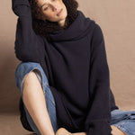 A woman sits wearing an oversized cashmere sweater in a grey blue colour with jeans