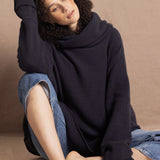 A woman sits wearing an oversized cashmere sweater in a grey blue colour with jeans