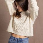 a woman wears a cropped cashmere aran sweater with her hands up in the air and wearing jeans
