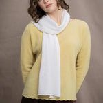 A woman wears a cashmere sweater jumper in pale yellow with scallop trim detail along neck, sleeves and hemline with brown silk trousers and white cashmere scarf on top