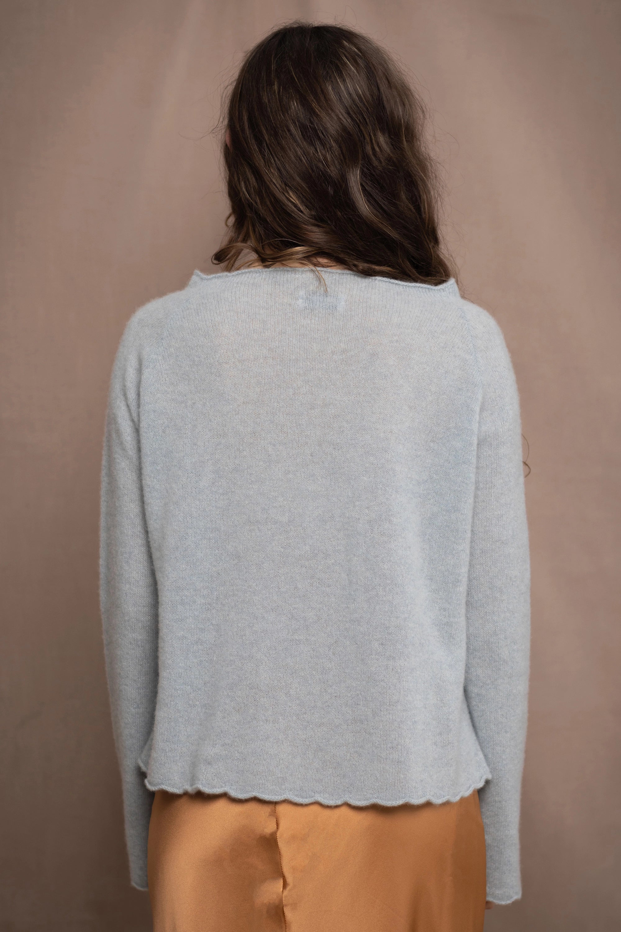 A woman wears a cashmere sweater jumper in pale turquoise blue with scallop trim detail along neck, sleeves and hemline