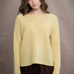 A woman wears a cashmere sweater jumper in pale yellow with scallop trim detail along neck, sleeves and hemline with brown silk trousers