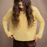 A woman wears a cashmere sweater jumper in pale yellow with scallop trim detail along neck, sleeves and hemline with brown silk trousers