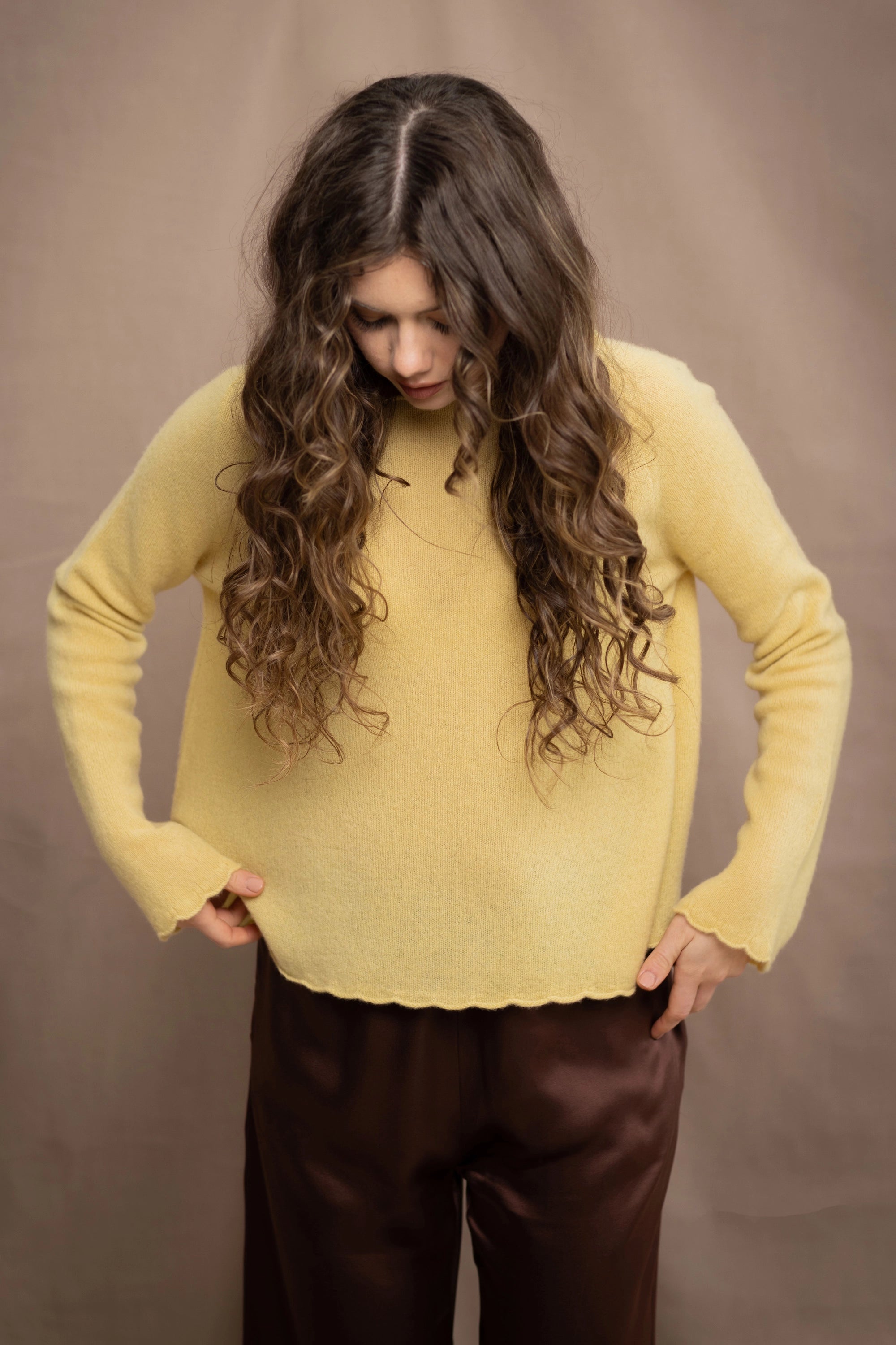 A woman wears a cashmere sweater jumper in pale yellow with scallop trim detail along neck, sleeves and hemline with brown silk trousers
