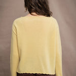 A woman wears a cashmere sweater jumper in pale yellow with scallop trim detail along neck, sleeves and hemline with brown silk trousers