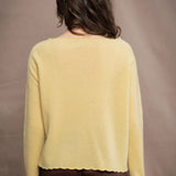 A woman wears a cashmere sweater jumper in pale yellow with scallop trim detail along neck, sleeves and hemline with brown silk trousers