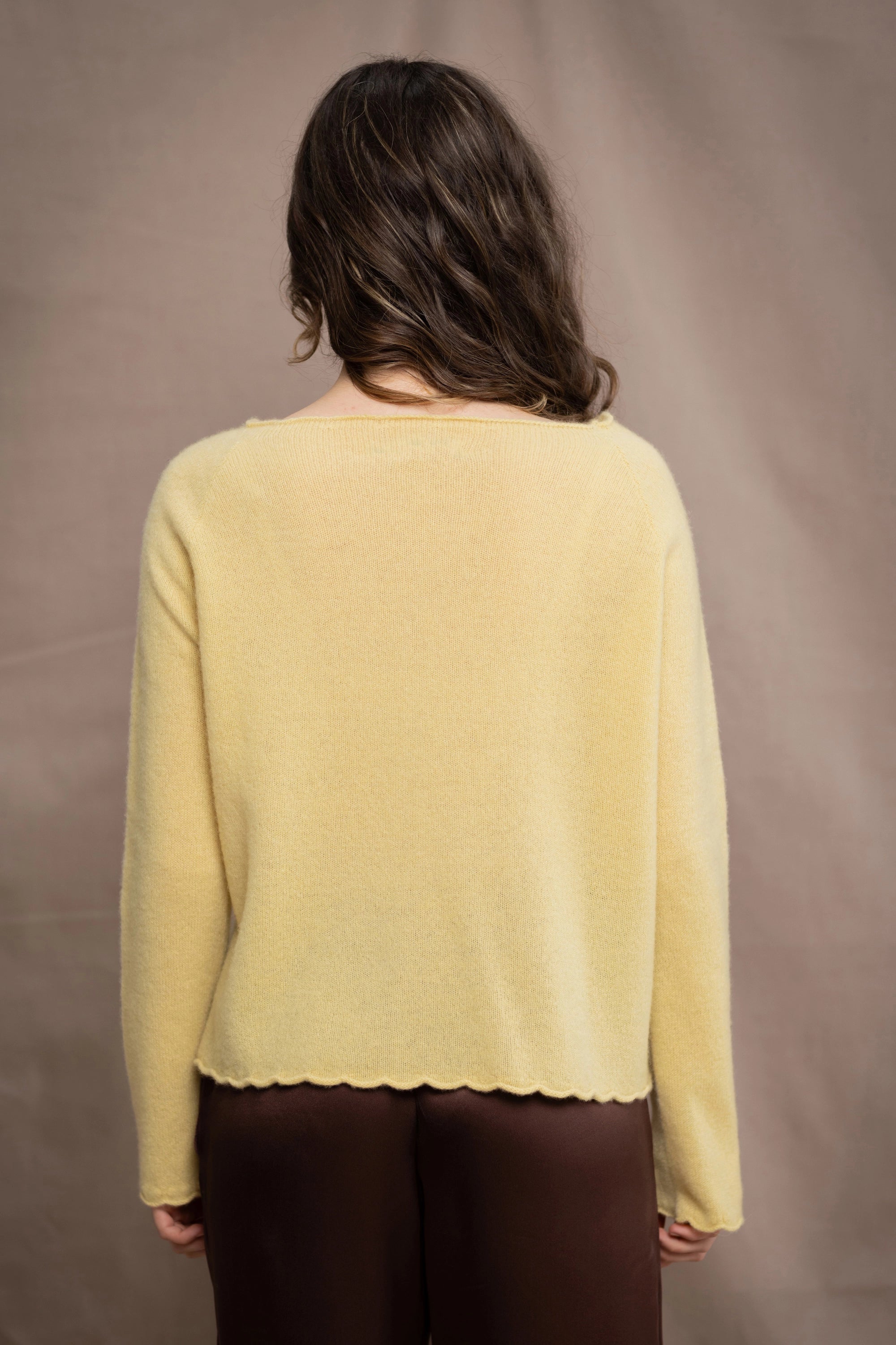 A woman wears a cashmere sweater jumper in pale yellow with scallop trim detail along neck, sleeves and hemline with brown silk trousers