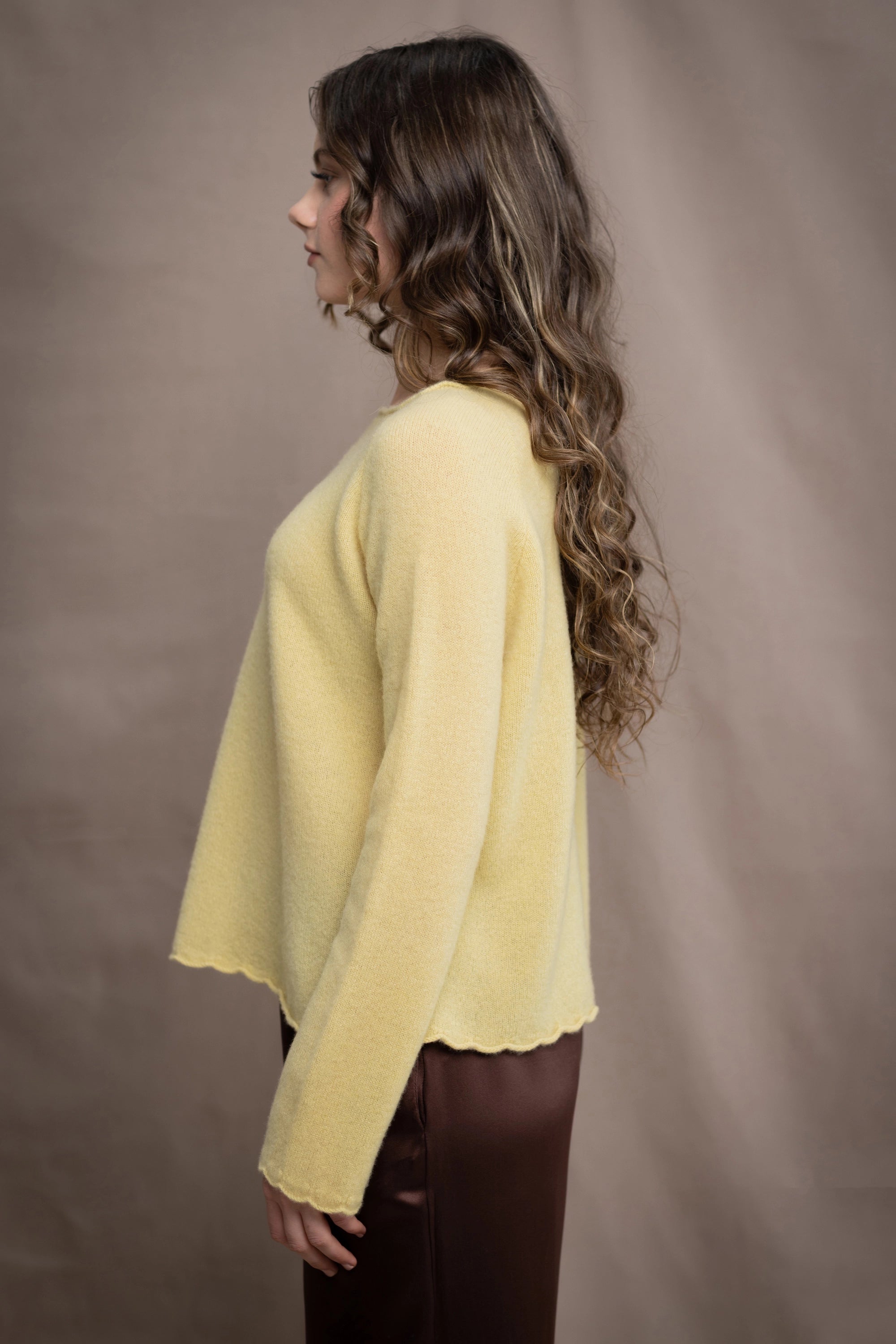A woman wears a cashmere sweater jumper in pale yellow with scallop trim detail along neck, sleeves and hemline with brown silk trousers