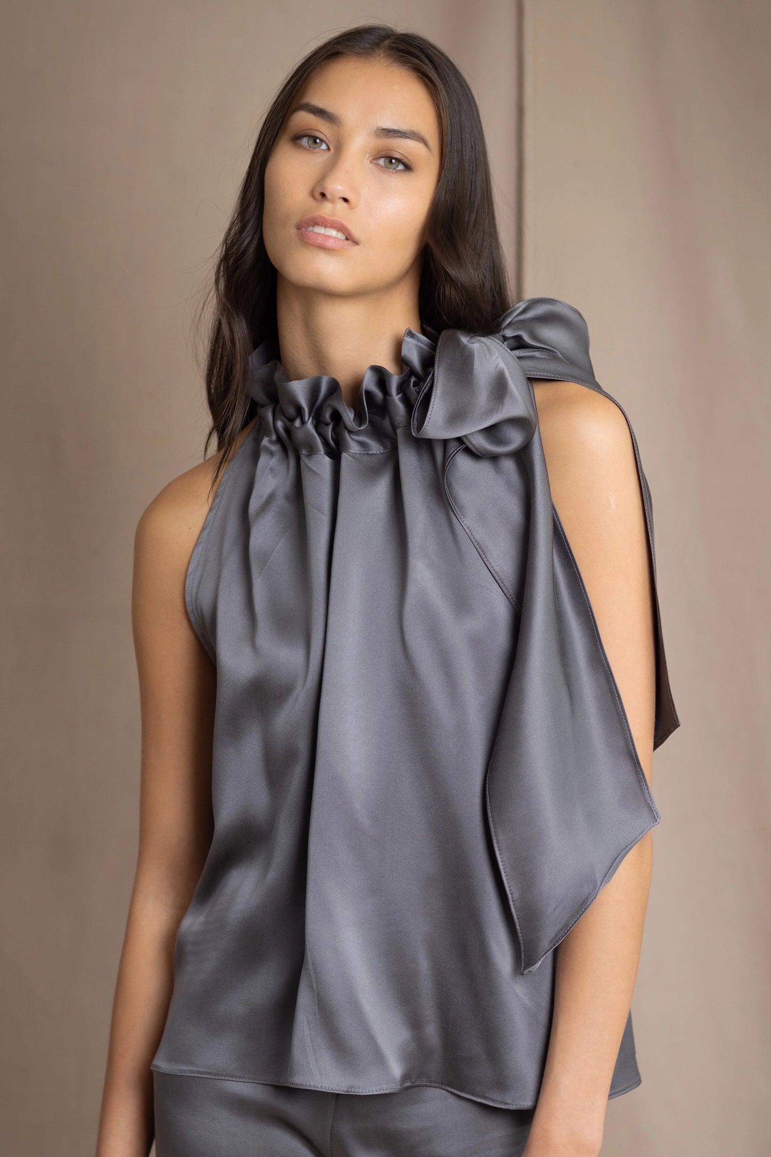A woman wears  a sleeveless silk top in grey with a dramatic ruffled high neckline  bow paired with matching gray silk pants