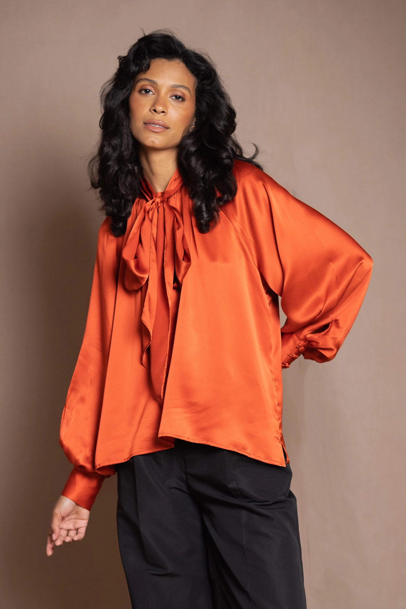 A woman modeling a vibrant orange silk blouse with a large bow tie neckline and voluminous balloon sleeves, paired with black trousers by Irish luxury brand Madigan Cashmere