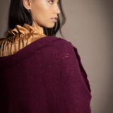 A woman wears a deep plum burgundy textured cashmere knit shawl draped over gold silk clothing by Irish brand Madigan Cashmere 