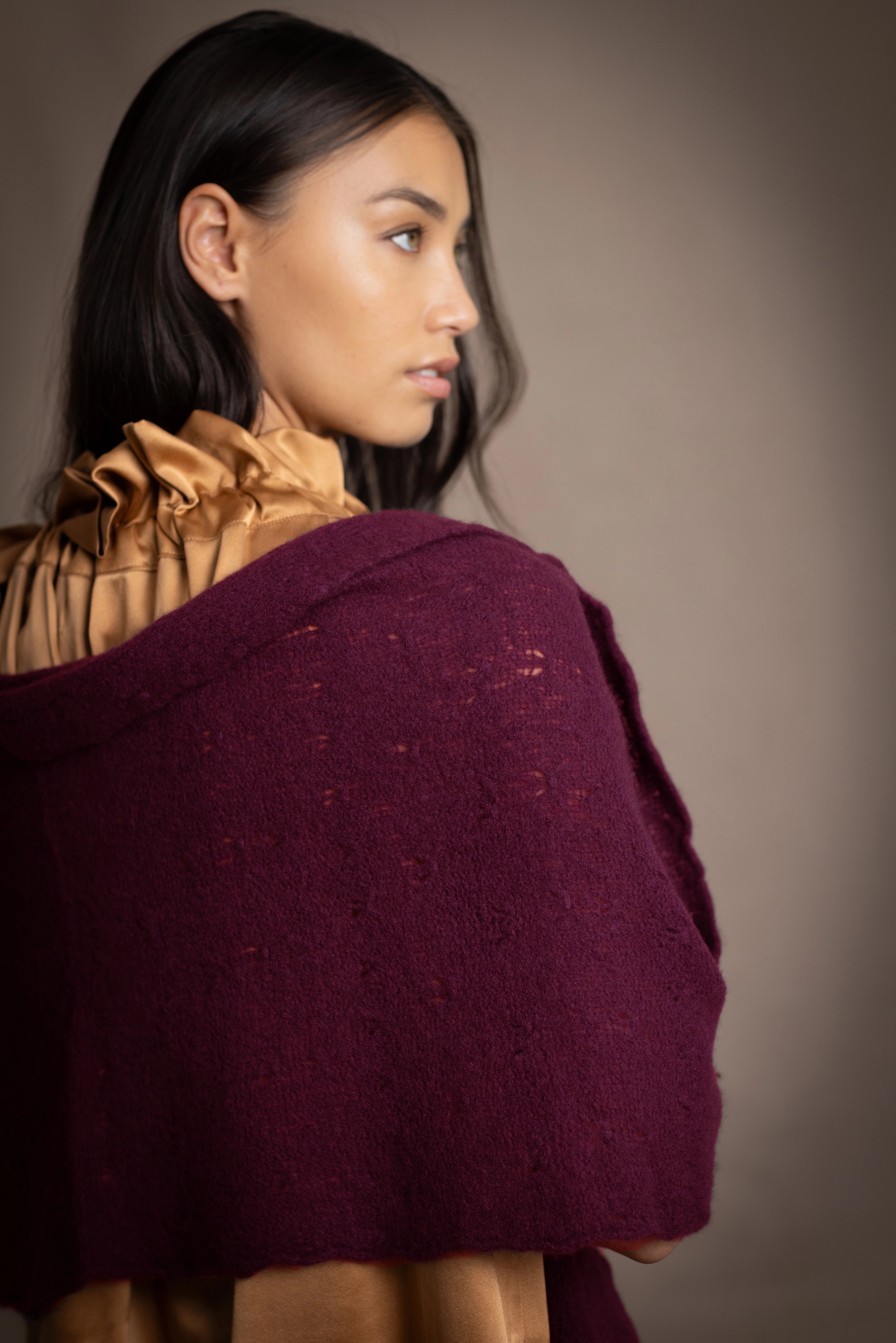 A woman wears a deep plum burgundy textured cashmere knit shawl draped over gold silk clothing by Irish brand Madigan Cashmere 
