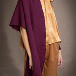 A woman wears a deep plum burgundy textured cashmere knit shawl draped over gold silk clothing by Irish brand Madigan Cashmere 