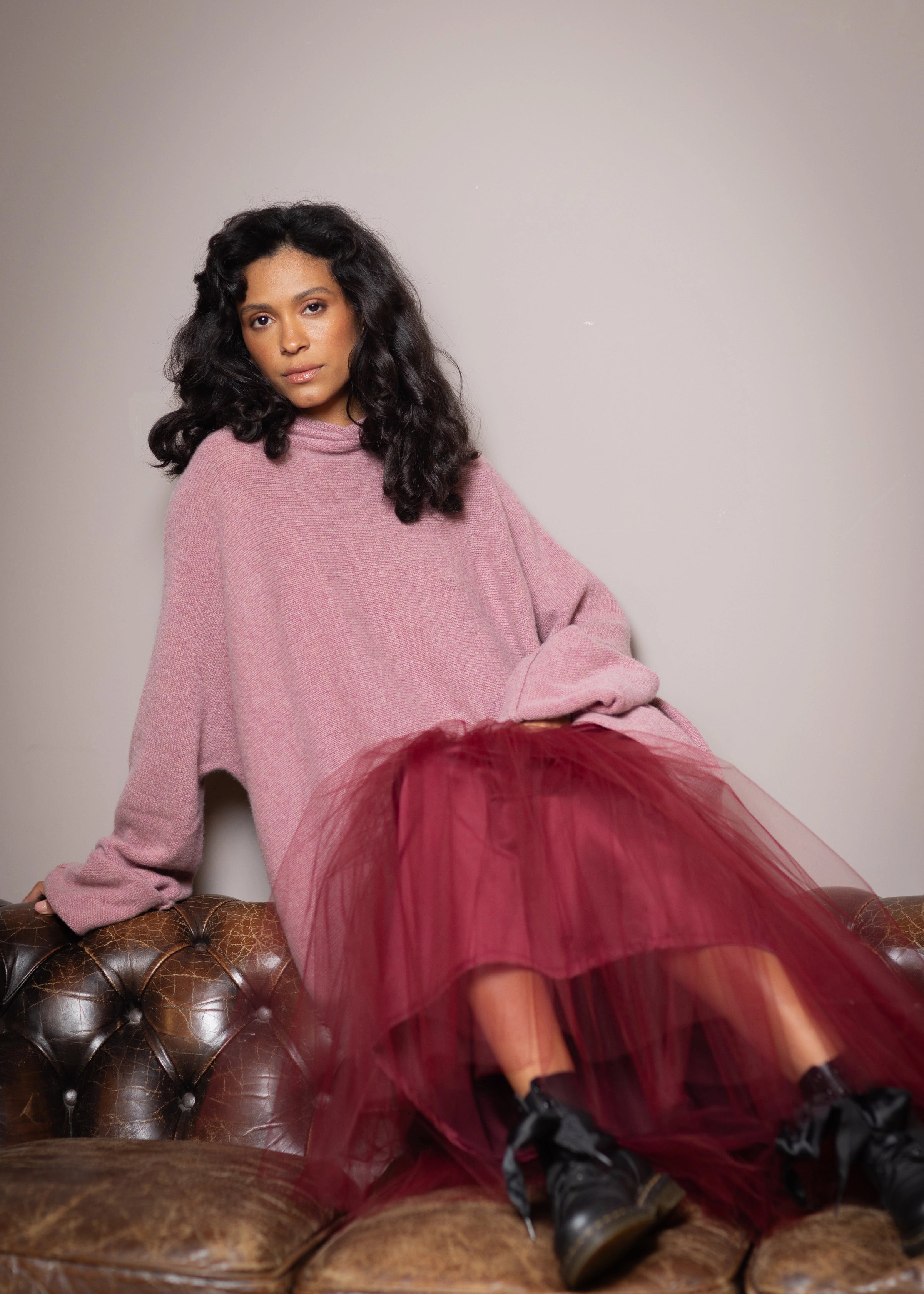 A woman wearing an oversized dusty pink knit cashmere sweater and a layered burgundy tulle skirt with black lace-up boots sits on a brown leather tufted couch 