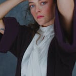 A woman wears a 100% silk pussybow sleeveless blouse in white with a brown and purple cashmere coat over top