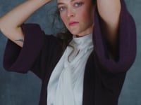 A woman wears a 100% silk pussybow sleeveless blouse in white with a brown and purple cashmere coat over top